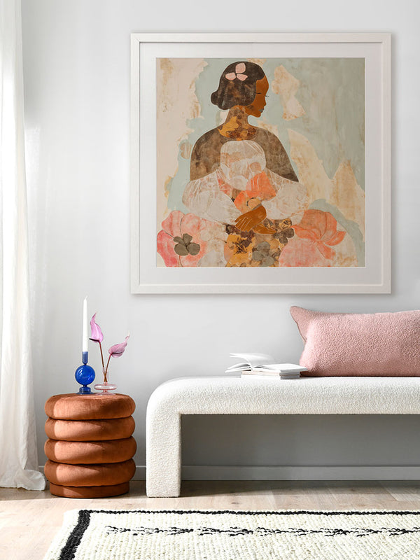 A framed Dreaming of Marigolds art print hanging in a modern neutral-toned living room. The print features a woman in warm floral tones, styled with a beige sofa, textured pillows, and a white coffee table.