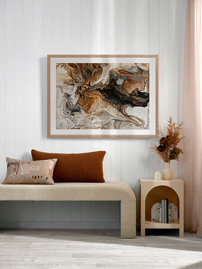 A framed Terrain Tides I art print hanging above a light-toned bench in a modern living room. The abstract artwork features swirling brown, black, and beige tones, complementing neutral decor.