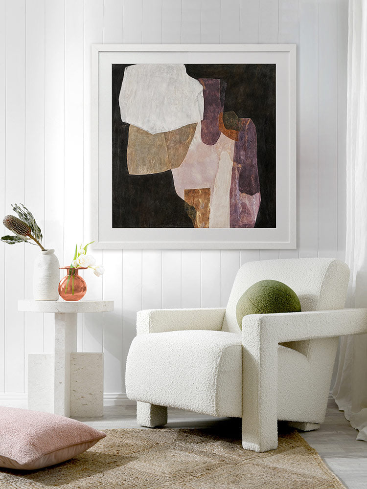 A framed Chiaroscuro I art print displayed above a white chair in a light, modern living space. The artwork's warm, earthy hues complement the bright decor.