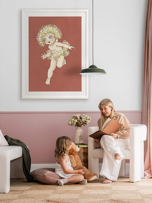 A woman reads to a girl with a teddy bear in a cozy, pink-accented room. Above them is Urban Road's Gum Blossom Baby I Terracotta Framed Art Print, featuring whimsical Australian flora. Flowers decorate the nearby table.