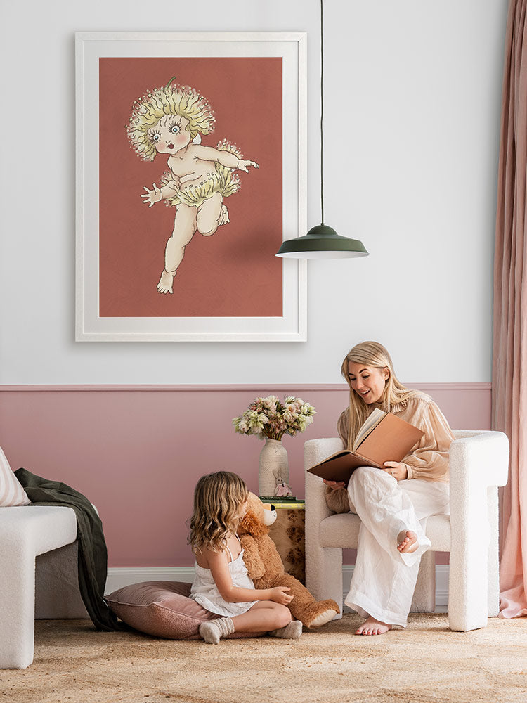 A woman reads to a girl with a teddy bear in a cozy, pink-accented room. Above them is Urban Road's Gum Blossom Baby I Terracotta Framed Art Print, featuring whimsical Australian flora. Flowers decorate the nearby table.