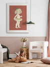 A young girl smiles on a soft rug with her teddy and book. Behind her, Urban Road's Gum Blossom Baby II Terracotta Framed Art Print hangs on a pink wall above a modern white chair.