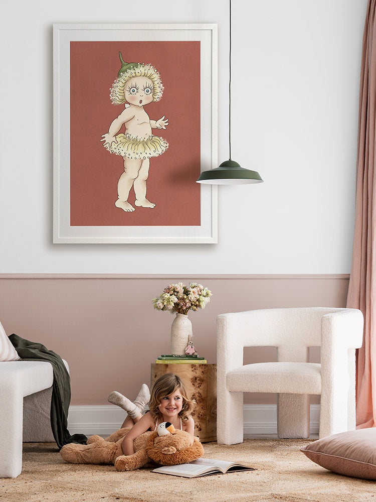 A young girl smiles on a soft rug with her teddy and book. Behind her, Urban Road's Gum Blossom Baby II Terracotta Framed Art Print hangs on a pink wall above a modern white chair.