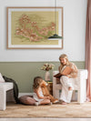 A woman reads to a young girl holding a teddy bear as they sit on a white armchair in a stylish living room. The green and white walls are decorated with "Picnic by the Billabong" framed art print by Urban Road and a vase of flowers.