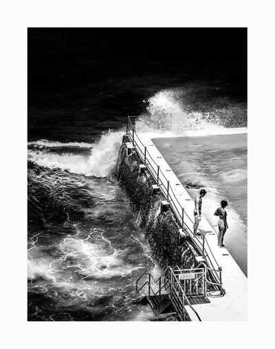 The Icebergs Unframed Art Print by Urban Road features a monochromatic photo of two people at Bondi Icebergs, standing by a seaside pool as waves crash and water splashes onto the deck.