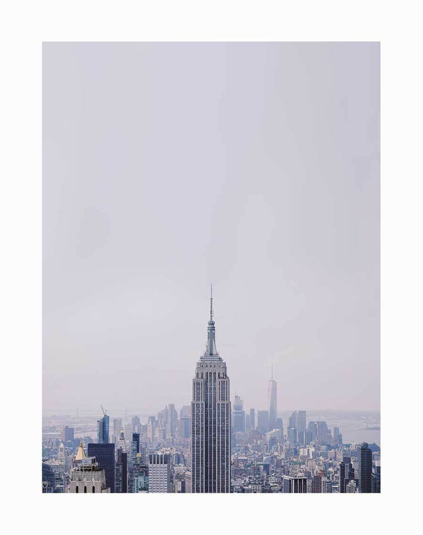 The Urban Road Rockefeller Unframed Art Print features a scenic view of New York City at dusk, with the Empire State Building centered in the foreground and skyscrapers beneath a pale, cloudy sky.