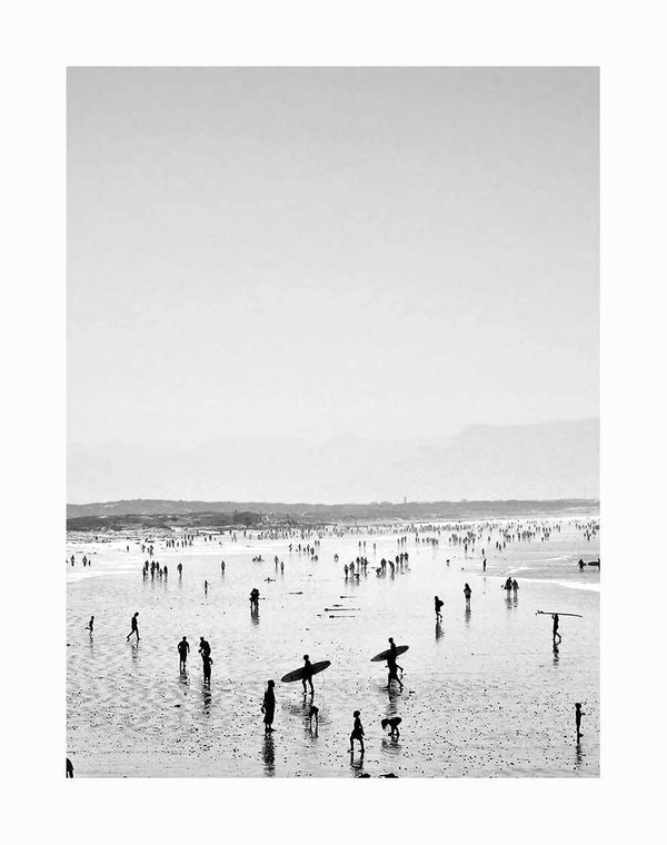 The Summer Wave I Unframed Art Print by Urban Road features black and white beach photography with strolling crowds, surfers carrying boards on wet sand with mirrored silhouettes, and a distant shoreline fading into a hazy sky.