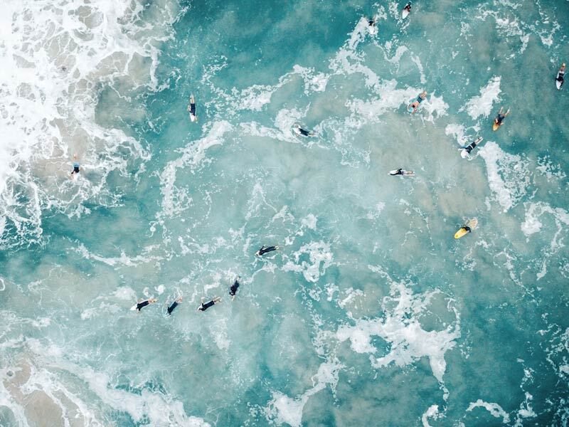 Urban Road's Surf's Up Unframed Art Print captures aerial surfers scattered in a clear blue sea near breaking waves, with foamy white surf visible in the top left corner.