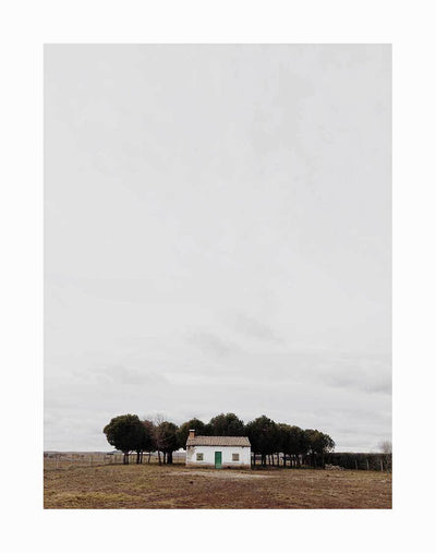 The Little Cottage Unframed Art Print by Urban Road features a small white cottage with a green door and tiled roof, set on a brown field before trees and a cloudy sky.