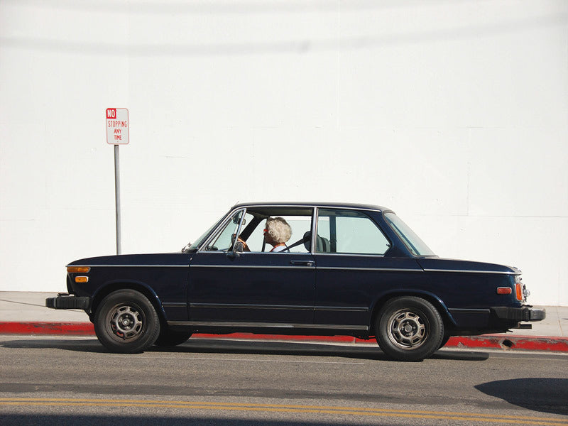 The Runaway Rhonda Unframed Art Print by Urban Road features an elderly woman at the wheel of a vintage black car parked by a red curb under a "No Parking Any Time" sign, creating a humorous scene against a plain white wall.