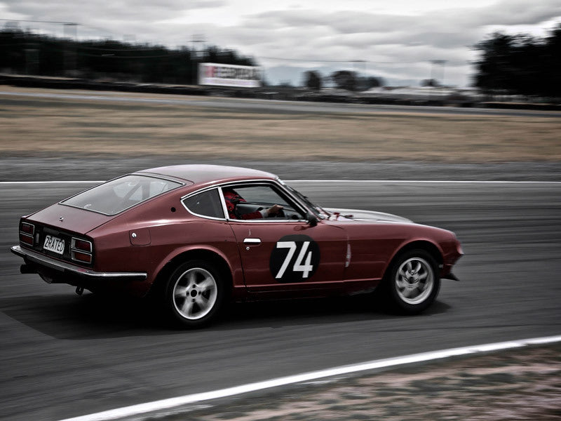 The Speed Racer Unframed Art Print by Urban Road captures a vintage red race car, number 74, racing around a track beneath overcast skies with blurred motion details that emphasize intensity and speed.