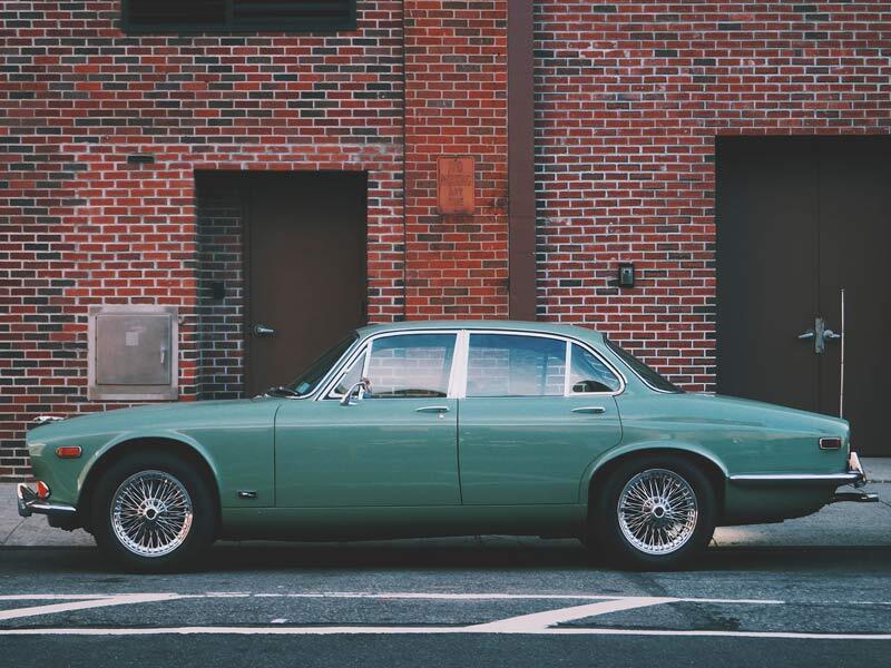 The Jag Unframed Art Print by Urban Road showcases a green vintage sedan with chrome features, wire-spoke wheels, and white street lines parked beside a red brick building with black doors and windows.