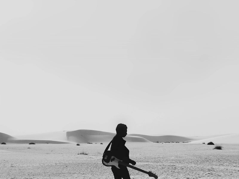 The Boss Unframed Art Print by Urban Road features a man with his guitar crossing a barren desert with distant dunes beneath a bright, hazy sky in striking black and white photography.