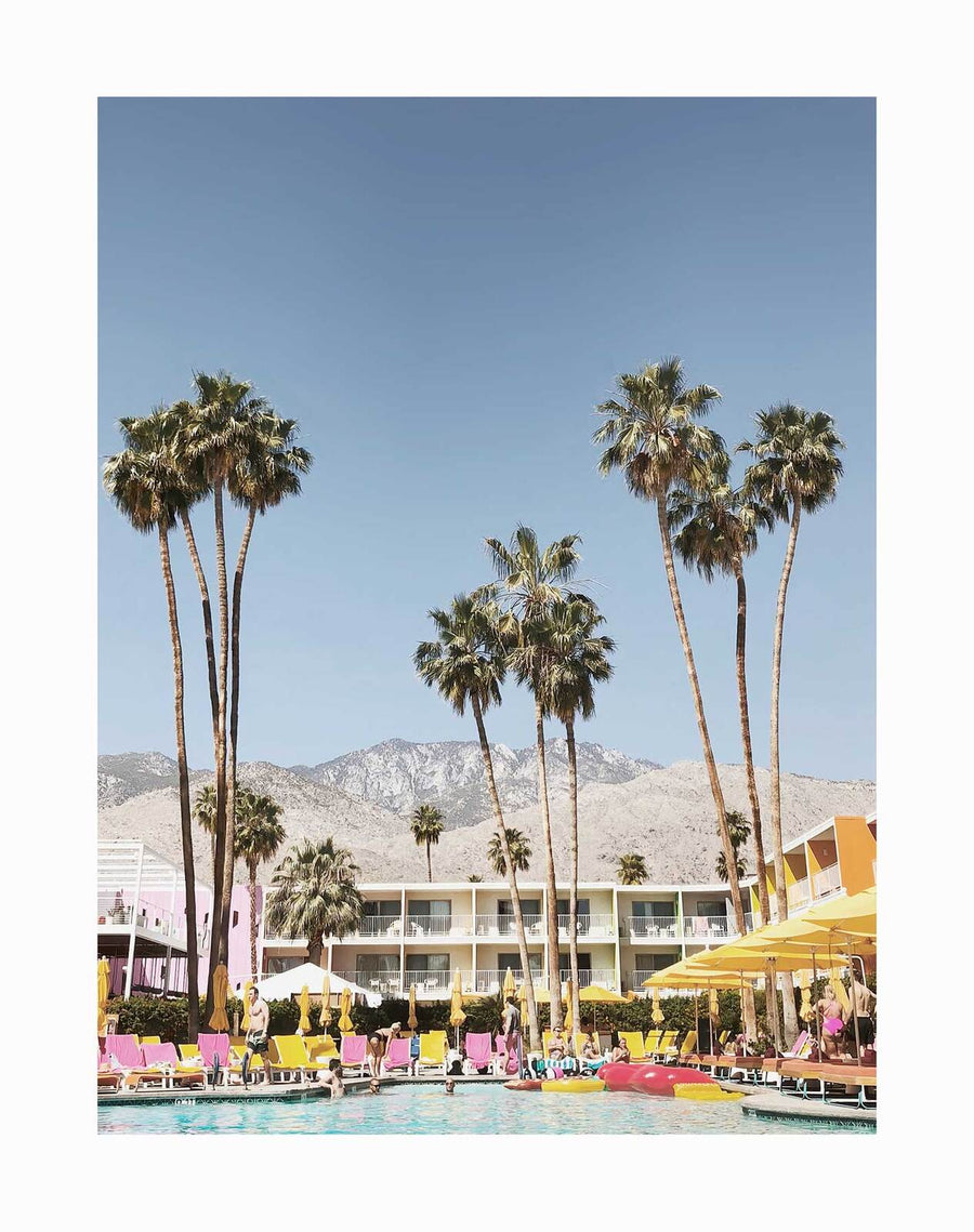 The Poolside at the Saguaro Unframed Art Print by Urban Road captures tall palms, yellow umbrellas, pink lounge chairs, and people relaxing by a vibrant pool at the Saguaro Hotel, set against mountains and a clear blue sky.