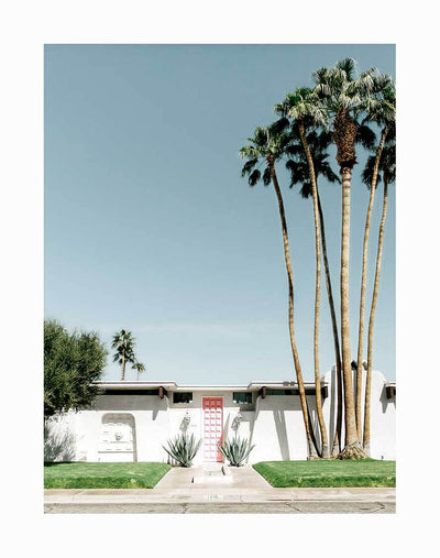 The "Sunrise Vista Unframed Art Print" by Urban Road features a mid-century modern white house with a pink door, lush grass, palm trees, and agave plants under a blue sky—capturing the essence of Palm Springs style.