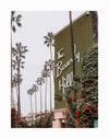 The Beverly Hills Hotel Unframed Art Print by Urban Road showcases a large green wall with "The Beverly Hills" in white script, framed by tall palms and lush greenery, and part of the hotel's iconic pink building in the background.