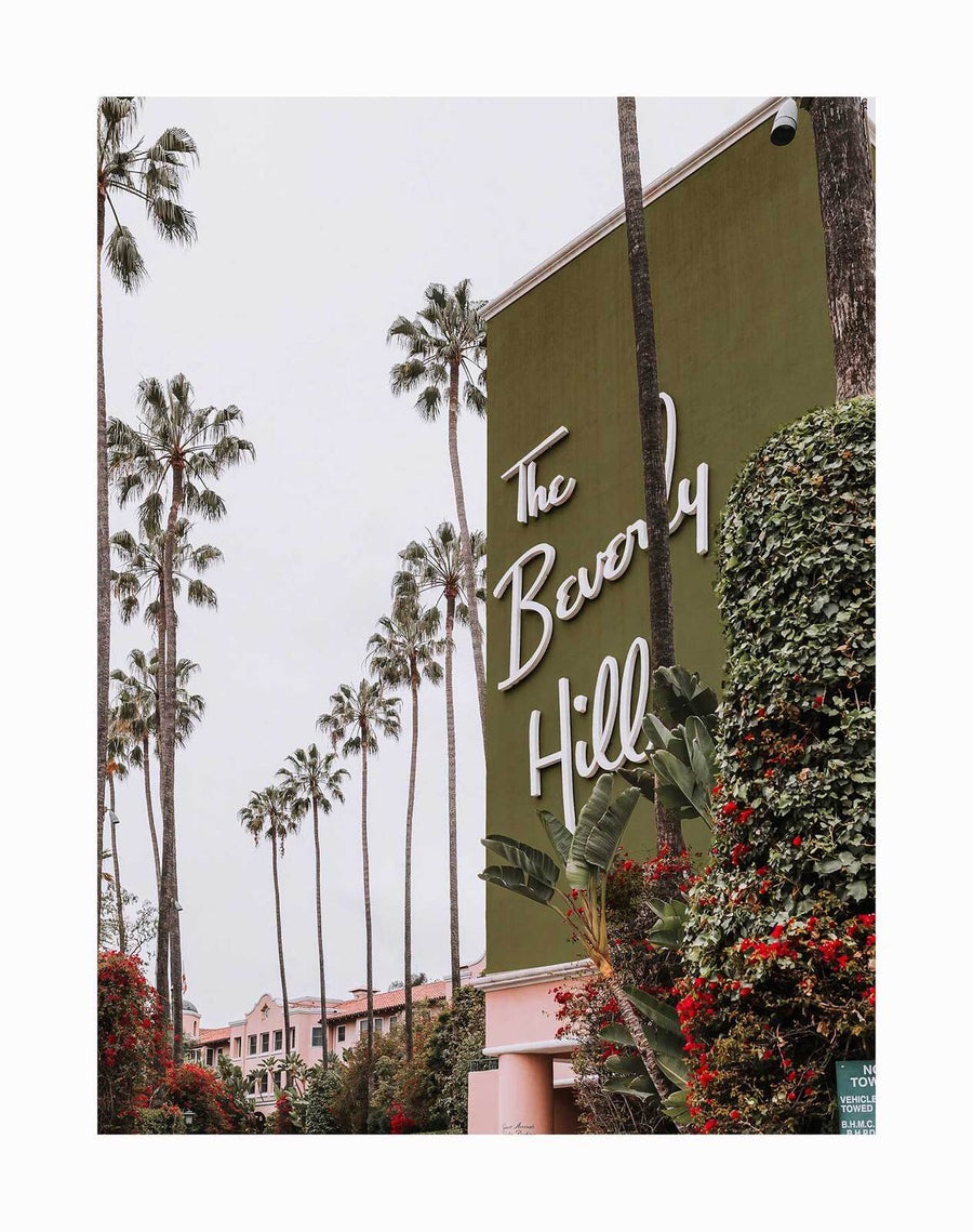 The Beverly Hills Hotel Unframed Art Print by Urban Road showcases a large green wall with "The Beverly Hills" in white script, framed by tall palms and lush greenery, and part of the hotel's iconic pink building in the background.