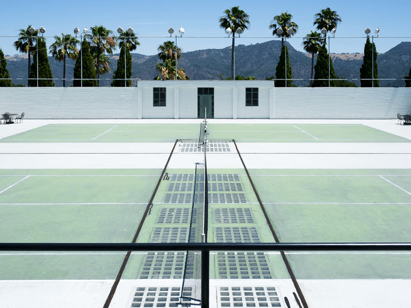 Down to the Tennis Court Unframed Art Print by Urban Road features empty outdoor tennis courts with light green surfaces, tall palm trees, California hills, a clear blue sky, and a white building in the background.