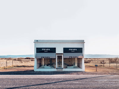 The Prada Unframed Art Print by Urban Road captures a small white Texas building with large display windows and three "PRADA MARFA" signs, standing alone in an open landscape with distant mountains under a pale sky.