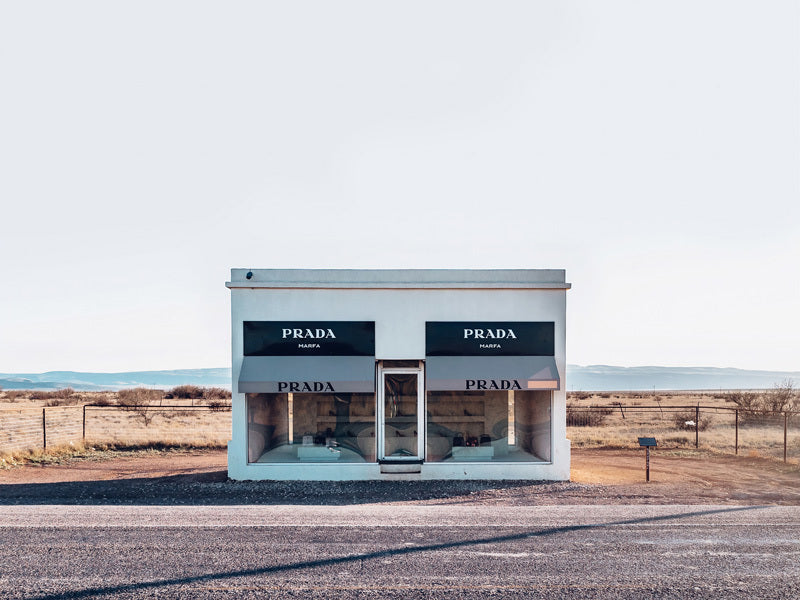 The Prada Unframed Art Print by Urban Road captures a small white Texas building with large display windows and three "PRADA MARFA" signs, standing alone in an open landscape with distant mountains under a pale sky.