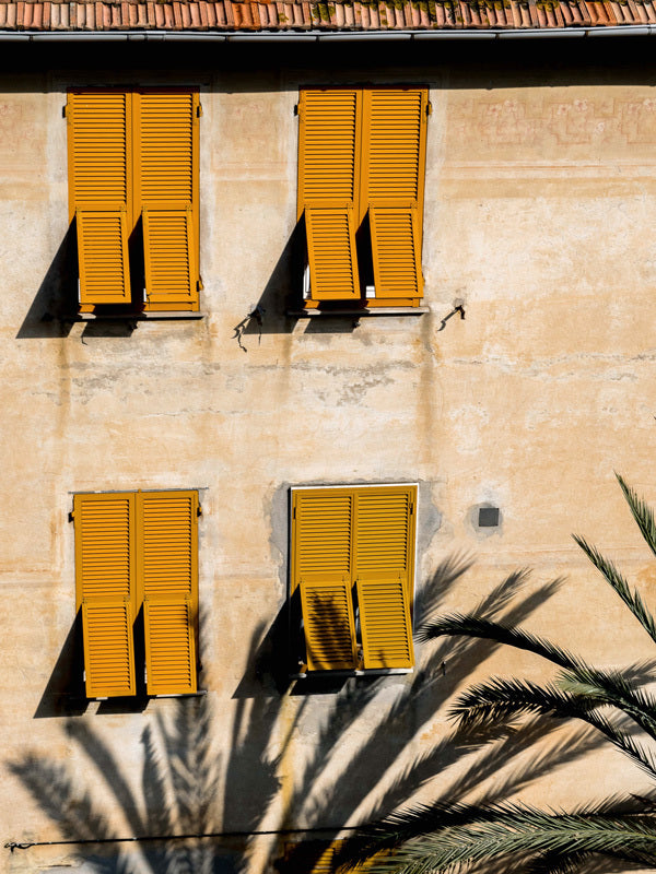 The Italian Clay Unframed Art Print by Urban Road features a beige wall with four yellow shutters, casting shadows as palm silhouettes stretch across this European facade in warm sunlight.