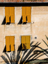 Four bright yellow shutters enliven a beige European facade, sunlight casting palm shadows over the windows in a scene captured by Urban Road’s Italian Clay Unframed Art Print.