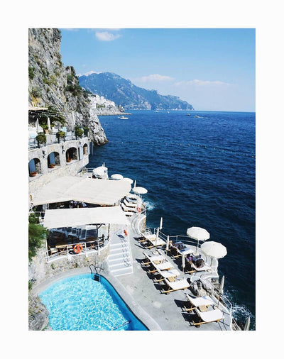 The Cliffs of Capri Unframed Art Print by Urban Road features a vibrant scene of an Italian cliffside resort with a pool, white umbrellas, and lounge chairs overlooking the dramatic Capri cliffs and deep blue ocean under a clear sky.