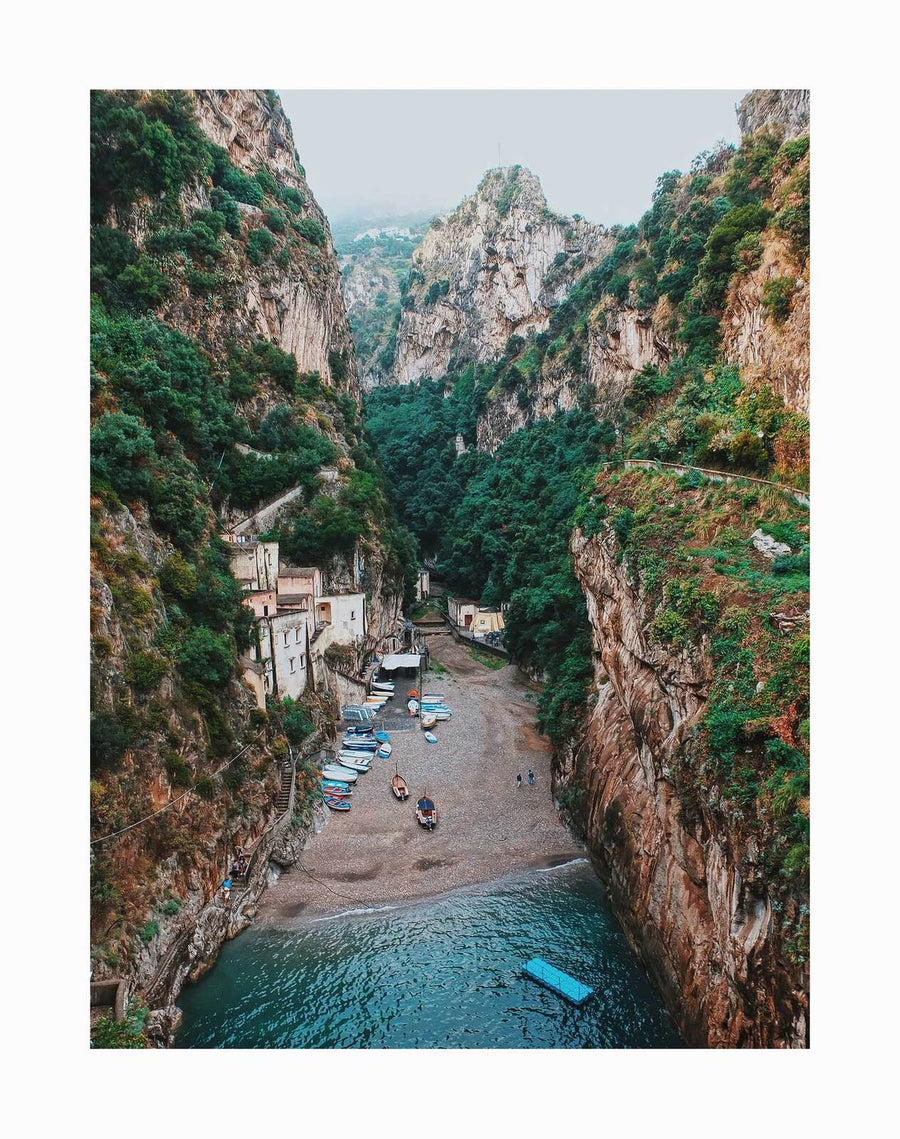 Discover the Mediterranean Adventure Unframed Art Print by Urban Road, featuring a hidden beach between rocky cliffs, lush greenery, small boats on the shore, and people relaxing by the clear sea—perfect for coastal-inspired decor.