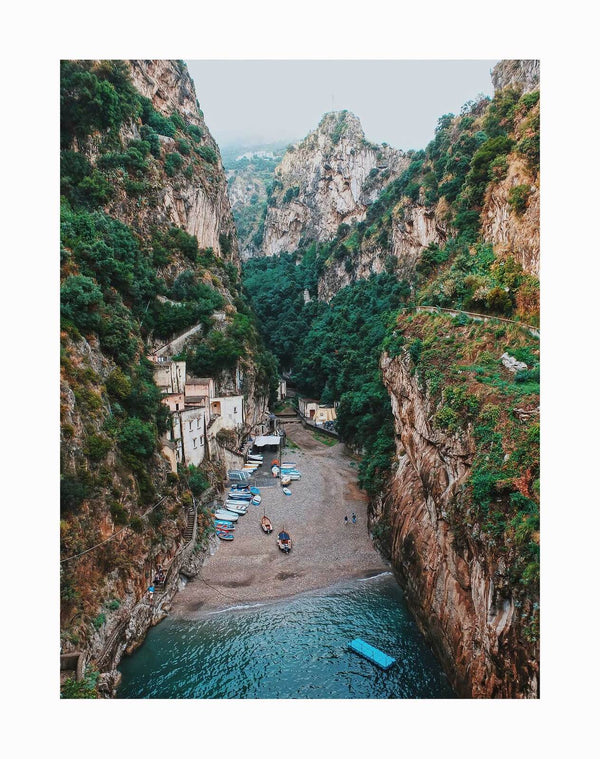 Discover the Mediterranean Adventure Unframed Art Print by Urban Road, featuring a hidden beach between rocky cliffs, lush greenery, small boats on the shore, and people relaxing by the clear sea—perfect for coastal-inspired decor.