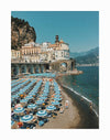 The Italian Beachgoers Unframed Art Print by Urban Road features a busy beach with blue umbrellas, pastel cliffside buildings, rocky coastlines, and mountains rising under a clear blue sky.