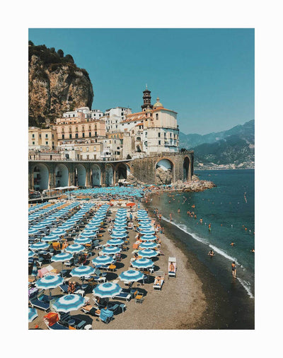 The Italian Beachgoers Unframed Art Print by Urban Road features a busy beach with blue umbrellas, pastel cliffside buildings, rocky coastlines, and mountains rising under a clear blue sky.