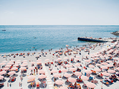 The Ciao Bella Unframed Art Print by Urban Road captures a classic Italian shoreline with red and white umbrellas, lounge chairs, sunbathers, swimmers, a blue sea, clear sky, and a pier—perfect for coastal decor.