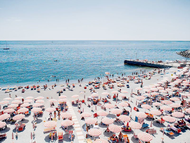 The Ciao Bella Unframed Art Print by Urban Road captures a classic Italian shoreline with red and white umbrellas, lounge chairs, sunbathers, swimmers, a blue sea, clear sky, and a pier—perfect for coastal decor.
