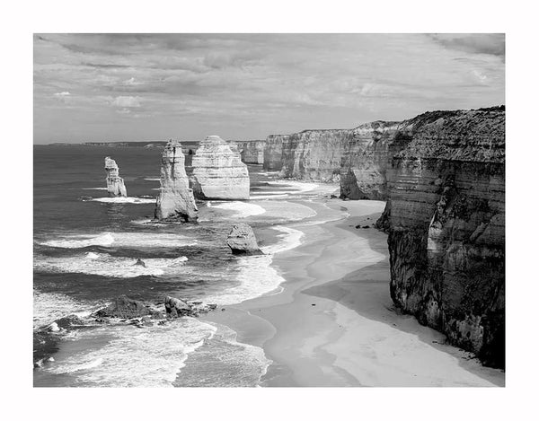 Twelve Apostles I Unframed Art Print by Urban Road features a black and white image of rugged sea cliffs and rock formations along Victoria’s coastline, with waves crashing on the beach under a partly cloudy sky.