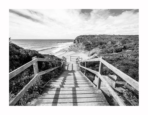 The Bells Beach I Unframed Art Print by Urban Road captures a wooden boardwalk leading to sandy shores and rolling waves, framed by lush greenery—ideal as a black and white poster for surfers and ocean lovers.