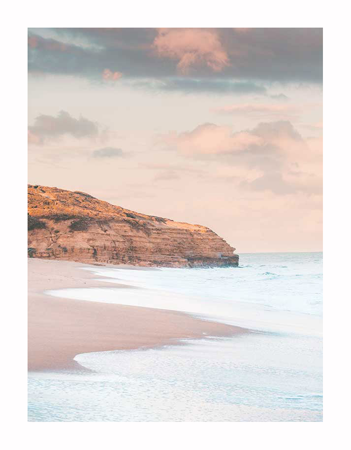 The Urban Road Sunset Beach Unframed Art Print captures gentle waves on sandy shores, a rocky cliff, and pastel clouds at sunset—an elegant photographic art piece that evokes calm and peace.