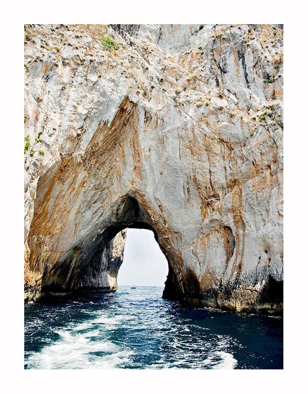 A natural rock arch rises above blue ocean waters, framed by rugged cliffs, in the stunning Grotto Unframed Art Print by Urban Road.
