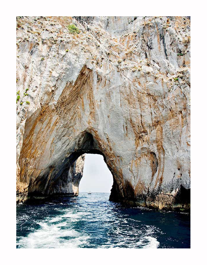 A natural rock arch rises above blue ocean waters, framed by rugged cliffs, in the stunning Grotto Unframed Art Print by Urban Road.