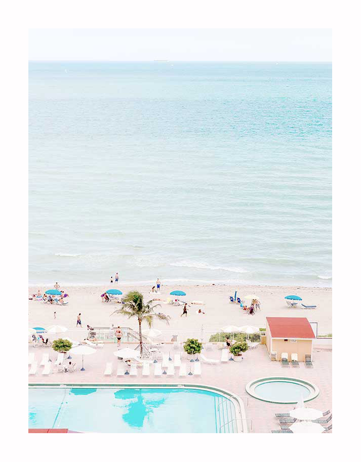 A bright, airy beach scene with colorful umbrellas, a calm turquoise ocean, and an empty pool with white lounge chairs—perfect as the Miami Poolside Unframed Art Print by Urban Road.