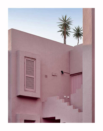 The Red Wall Unframed Art Print by Urban Road depicts a brownish-red geometric building with a closed shuttered window, stairs, and door. Two tall palm trees stand in the background beneath a clear blue sky.
