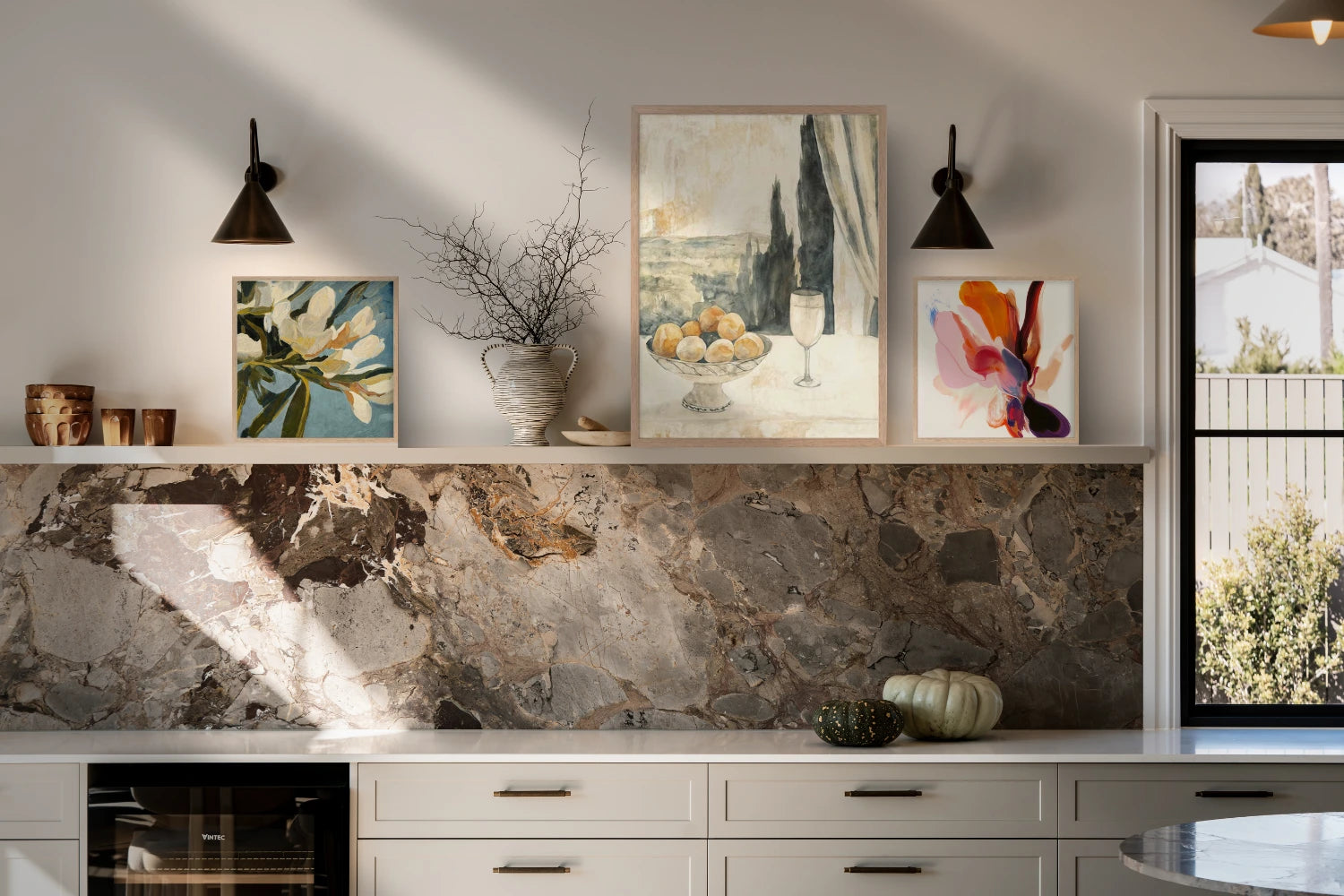 Kitchen shelf with art prints including a still life, floral painting, and colorful abstract piece above a marble backsplash.